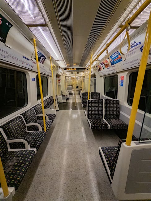 Interior view of an empty train carriage with a clean, polished grey floor and black fabric-covered seats featuring a multicoloured dotted pattern. Bright lighting from ceiling fixtures illuminates the beige ceiling with metallic ventilation panels and yellow handrails running along the sides. The windows are clear, and the space appears well-maintained and dust-free, reflecting professional cleaning standards. This scene showcases a neat environment suitable for surface cleaning and sanitisation, aligning with East Finchley rug cleaning tips near the tube station. Finchley Carpet Cleaners emphasizes hygiene and cleanliness in public transport settings, as seen in the image.