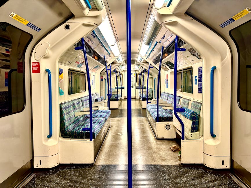 Interior of an empty London Underground train carriage with symmetrical seating arrangement on both sides. The seats are upholstered in multicoloured patterned fabric, appearing clean and well-maintained. The carriage has white plastic panels with blue handrails and poles for standing passengers. The flooring is dark, textured rubber, showing minimal debris, indicating recent cleaning or maintenance. Overhead, fluorescent lighting illuminates the space evenly, highlighting the spotless surfaces and the organized, hygienic condition of the carriage. Windows are visible along the sides, with some signage and safety notices displayed above. The overall scene emphasizes surface cleanliness and sanitation relevant to domestic cleaning or deep cleaning practices, as might be recommended by Finchley Carpet Cleaners for maintaining hygiene in shared public spaces or residential areas. The setting is well-lit and orderly, reflecting a focus on consistent cleanliness and surface sanitisation.
