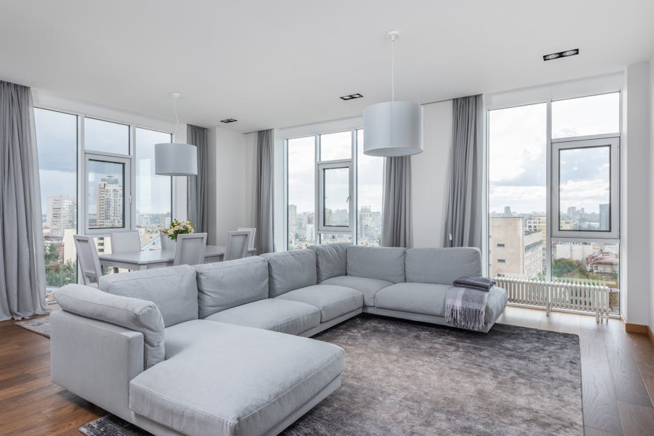 A spacious living room with large floor-to-ceiling windows providing a view of a cityscape, featuring a light grey L-shaped fabric sofa positioned on a dark grey rug. The room has white painted walls, and the hardwood floor is visible around the rug. Two white pendant lights hang from the ceiling, illuminating the room evenly. The windows are framed with light grey curtains, pulled back to let in natural light, highlighting the clean and tidy surface of the sofa and floor. No clutter or visual signs of dirt are visible, suggesting recent or ongoing surface cleaning and maintenance. The setting appears bright and freshly maintained, aligned with professional domestic cleaning standards by Finchley Carpet Cleaners.