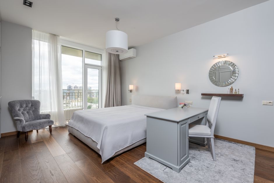 A bright, modern bedroom with a double bed covered in white bedding positioned against a white wall, flanked by two wall-mounted bedside lamps. To the left of the bed, a grey upholstered armchair with tufted detailing sits near large windows with sheer white curtains that allow natural light to fill the room. The windows offer a view of a city skyline with buildings in the distance. A sleek, white desk with a matching chair is placed at the foot of the bed, with a small stack of books on top. On the wall behind the desk, a round mirror with a metallic frame and a wall-mounted light above it add decorative detail. The room features a wooden floor and an air conditioning unit installed near the ceiling, creating a clean and hygienic environment suitable for routine surface and deep cleaning or domestic sanitisation by Finchley Carpet Cleaners as part of their residential cleaning services.