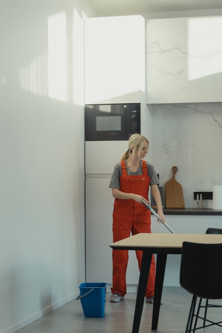 A woman with blonde hair wearing an orange apron and gray t-shirt is standing on a small step ladder, performing surface cleaning on a large, dark-colored wall in a modern, well-lit room. She is using