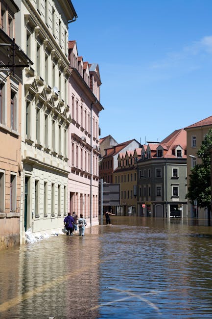 Photograph of a city street submerged in floodwater, with multi-story buildings featuring pastel-colored facades, white and beige window frames, and red-tiled roofs. The water covers the cobblestone p
